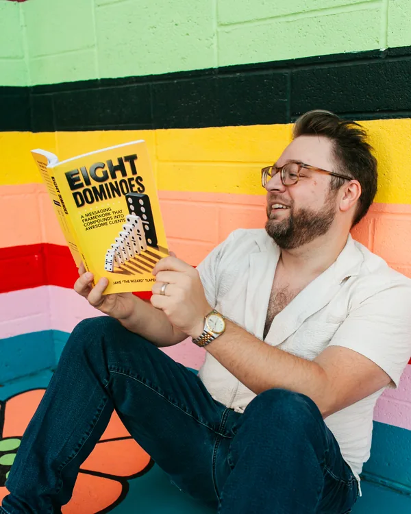 Jake Tlapek smiling down at a copy of Eight Dominoes in front of a striped mural