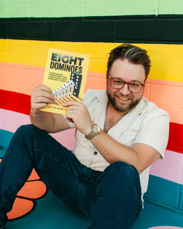 Jake "The Wizard" Tlapek smiling and holding up a copy of Eight Dominoes against a striped yellow wall