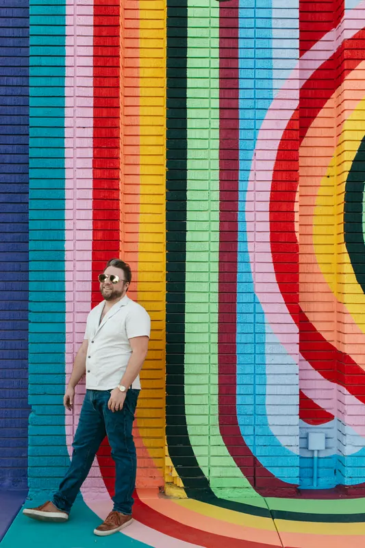 Jake Tlapek in sunglasses walking past a rainbow-striped brick wall
