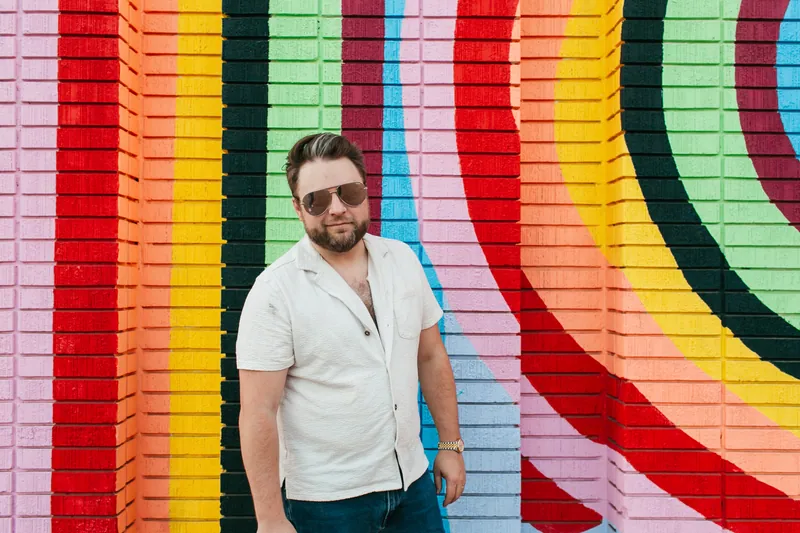Jake Tlapek in sunglasses, standing relaxed in front of a rainbow-striped brick mural