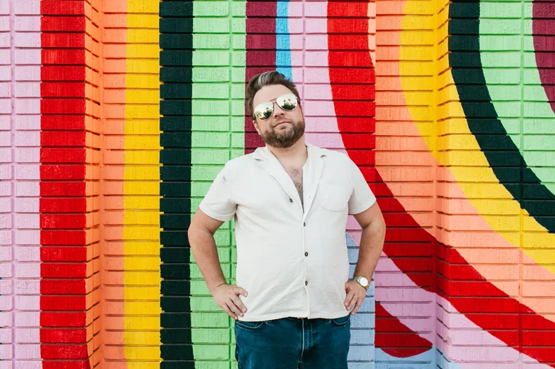 Jake Tlapek standing hands on hips in sunglasses in front of a rainbow-striped brick mural