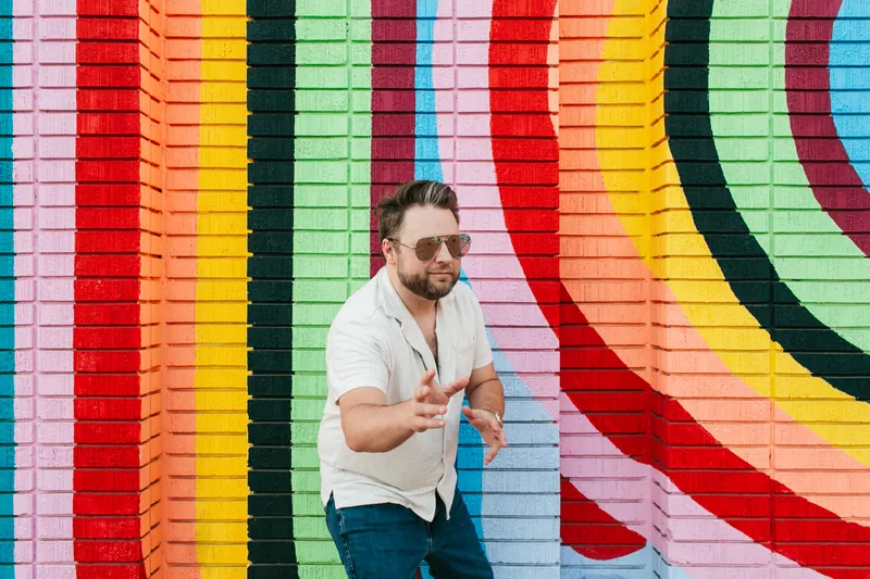 Jake Tlapek in sunglasses pointing both hands at the camera in front of a rainbow-striped brick mural
