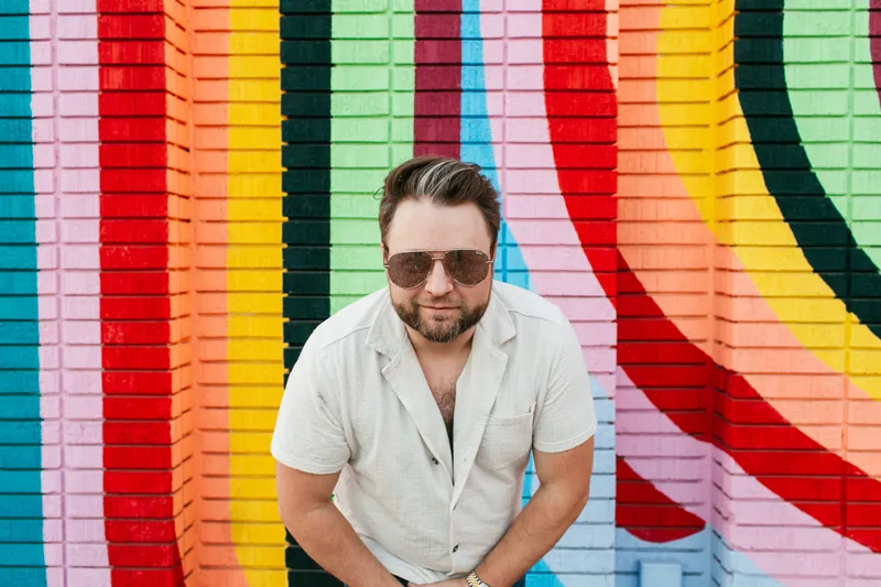 Jake Tlapek leaning in toward the camera in front of a rainbow mural