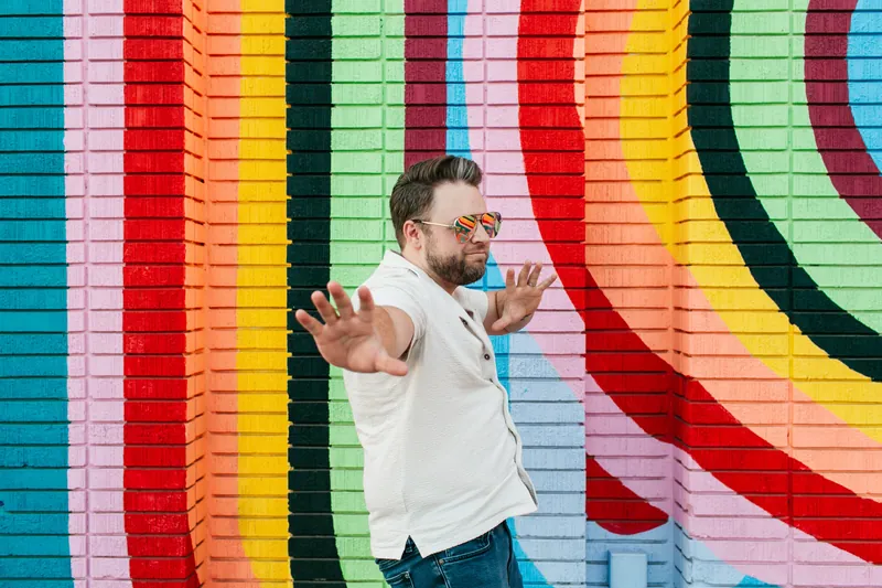 Jake Tlapek in sunglasses with his arms spread wide in welcome, in front of a rainbow-striped brick mural