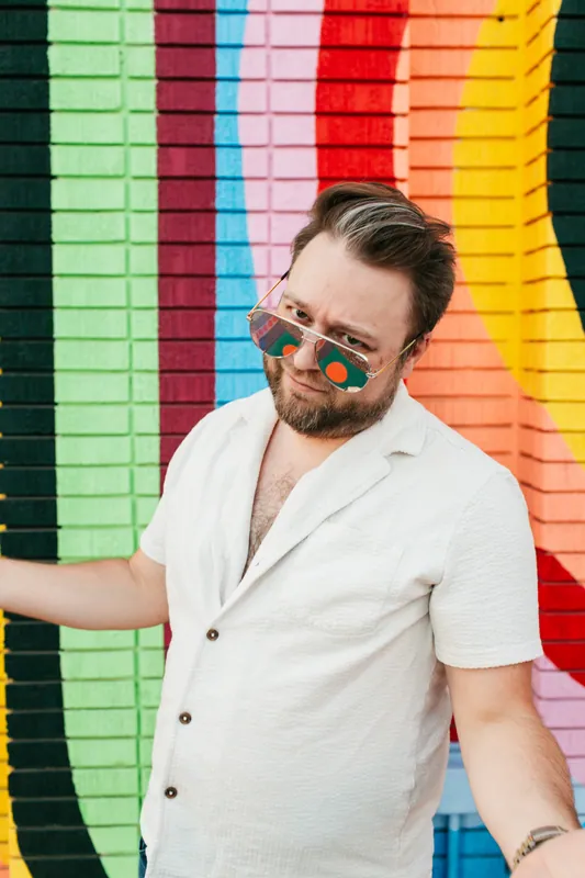 Jake Tlapek, head tilted, sunglasses on, leaning on a rainbow mural